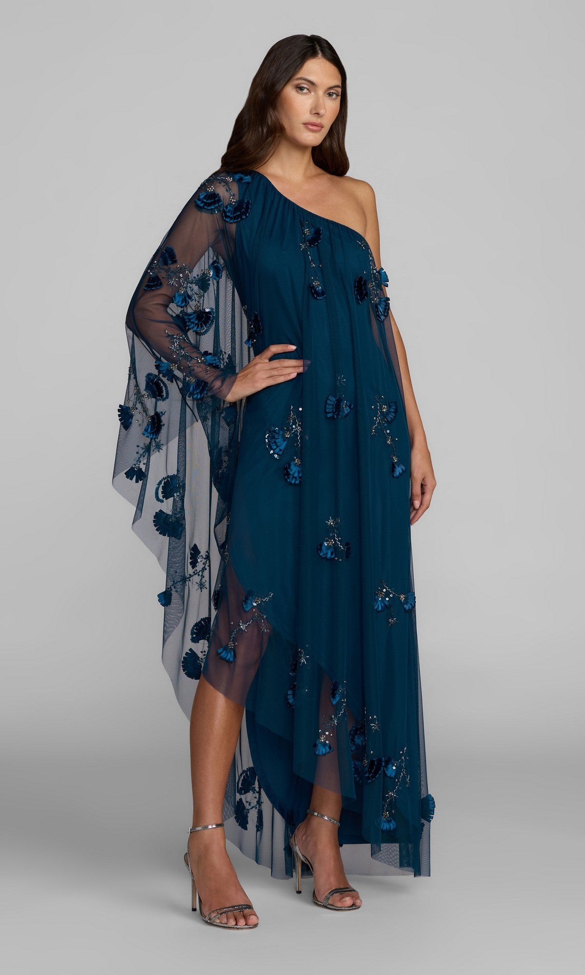 A woman stands against a plain gray background, wearing the Mac Duggal 9374 Long Formal Dress in twilight—a one-shoulder gown featuring floral appliqués, a sheer cape sleeve, and paired with silver high-heeled sandals for an elegant look.